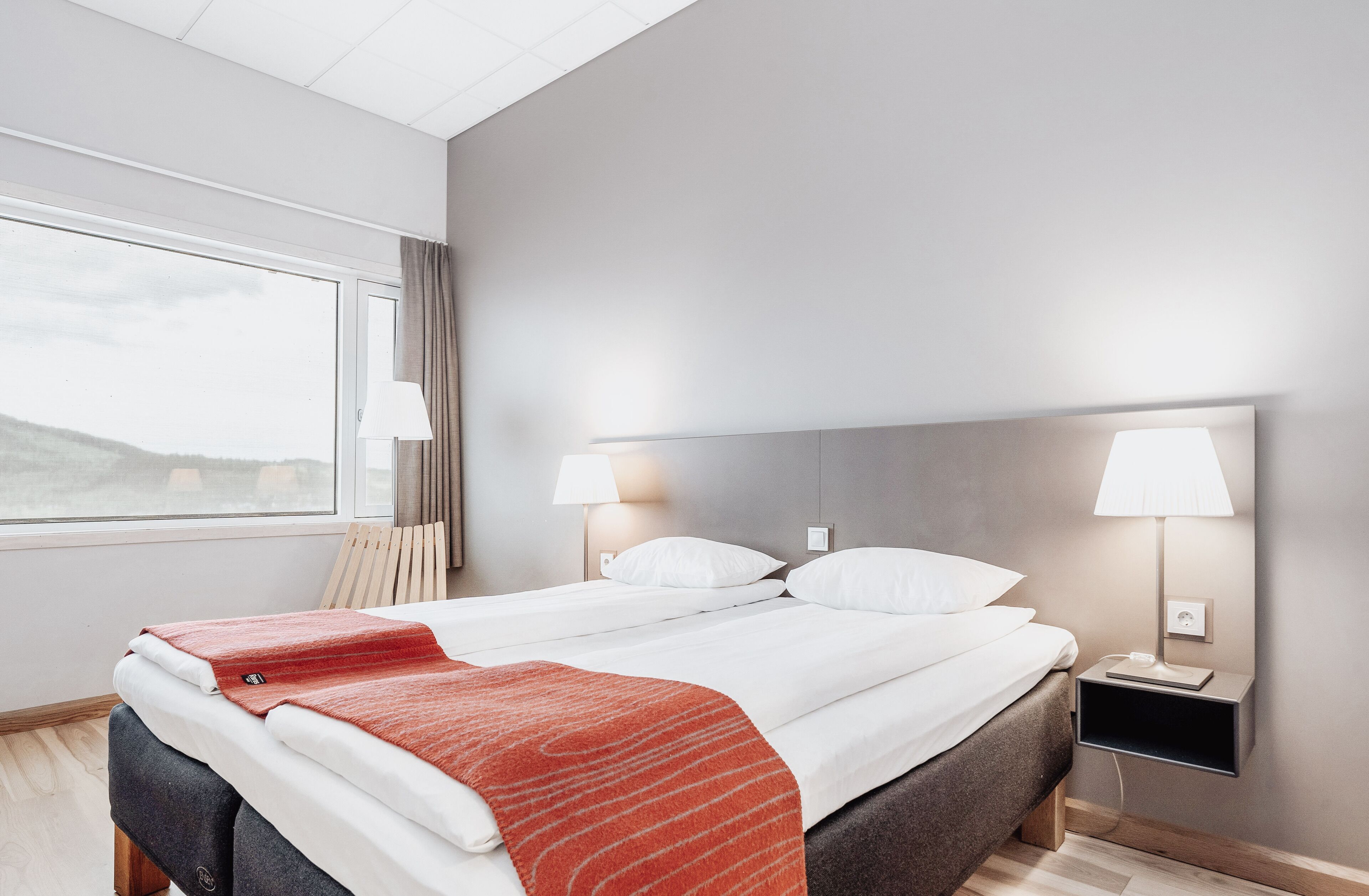 Quarto de hotel com cama, duas luminárias de cabeceira, mesa de cabeceira e janela com cortinas.