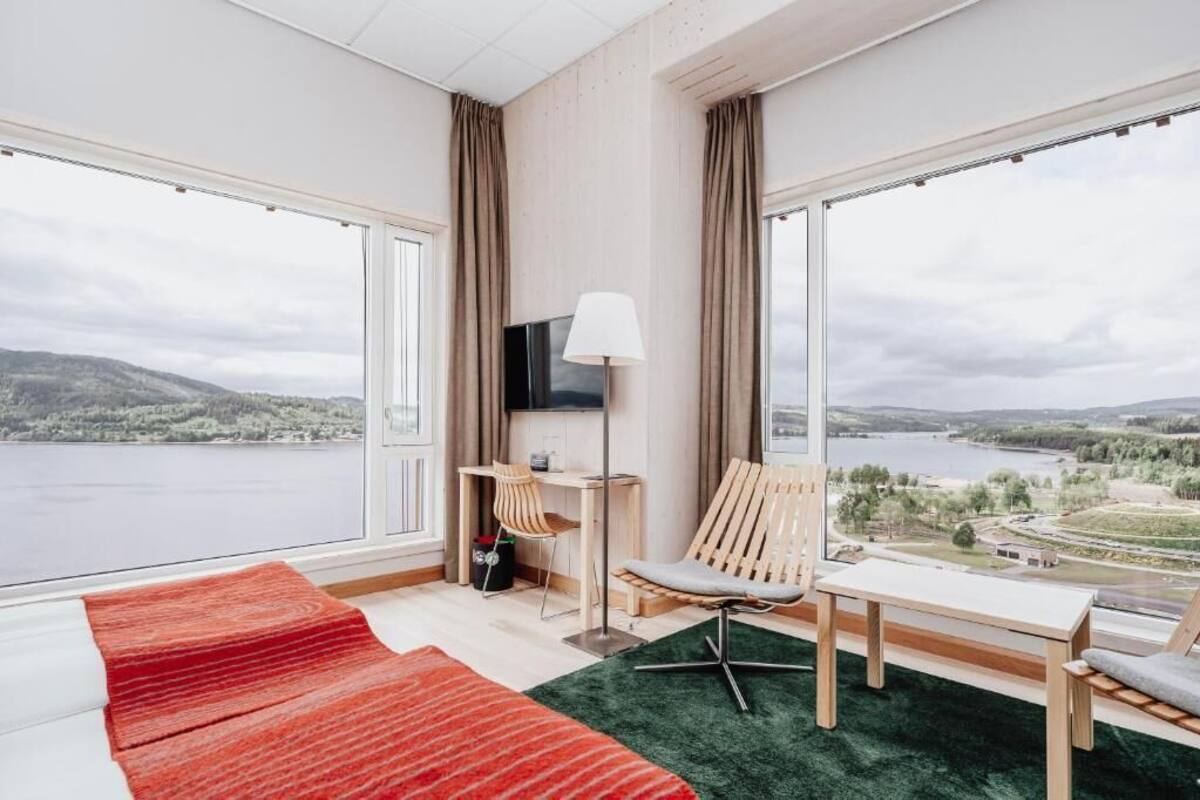 Habitación de hotel moderna con un ventanal grande que ofrece una vista panorámica de un lago y colinas. Se puede ver una cama, un escritorio con una silla y un televisor montado en la pared.