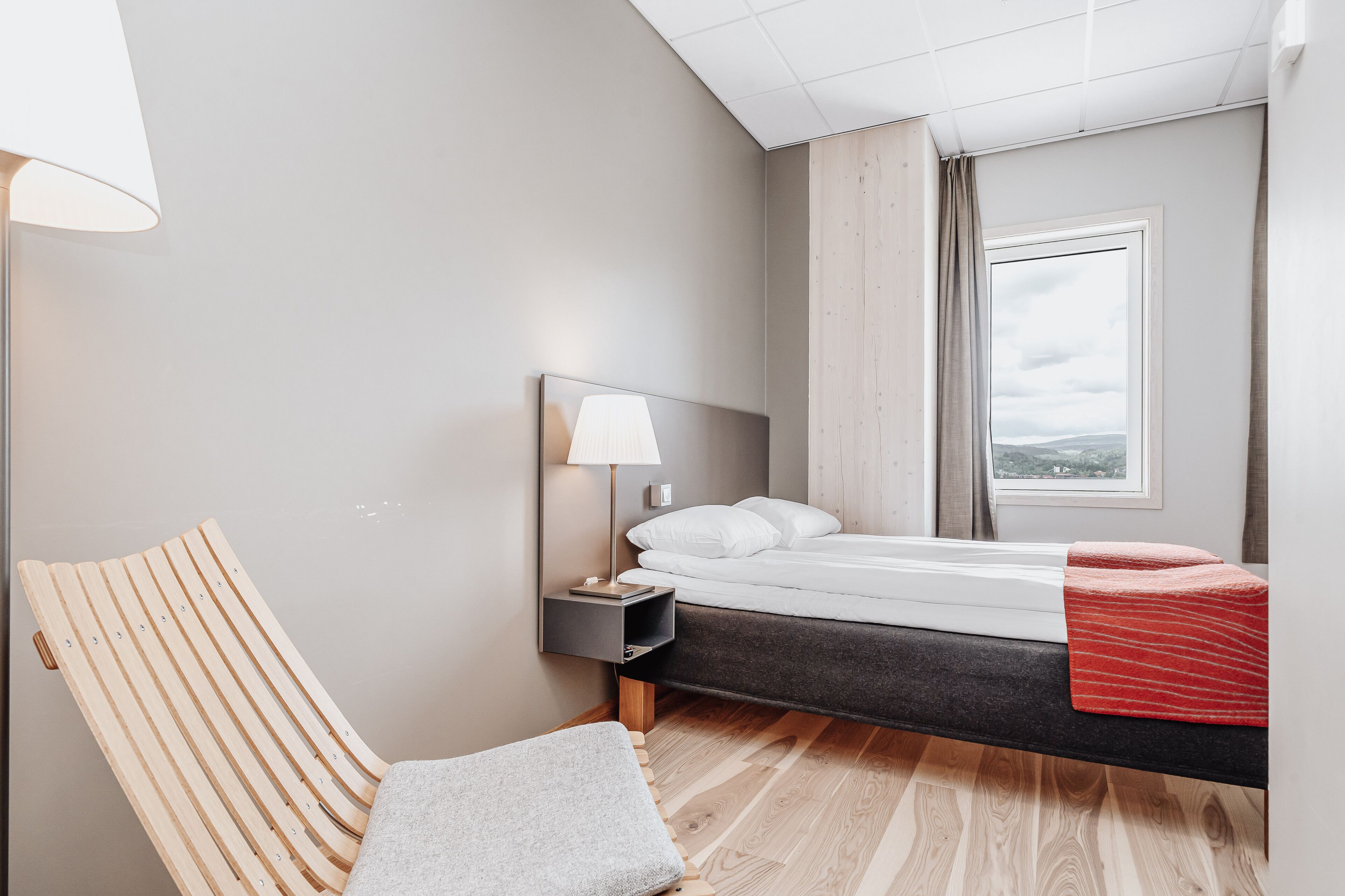 A modern hotel room with a bed, a nightstand with a lamp, a wooden chair, and a view of the outdoors.