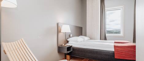 A modern hotel room with a bed, a nightstand with a lamp, a wooden chair, and a view of the outdoors.