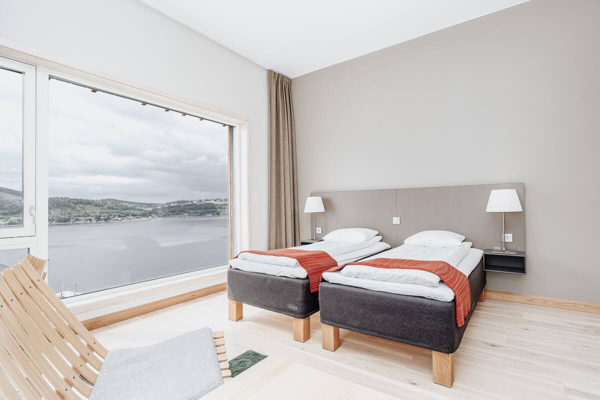 Una habitación de hotel moderna con dos camas, un ventanal con vistas a un lago, una silla de madera y una mesita.