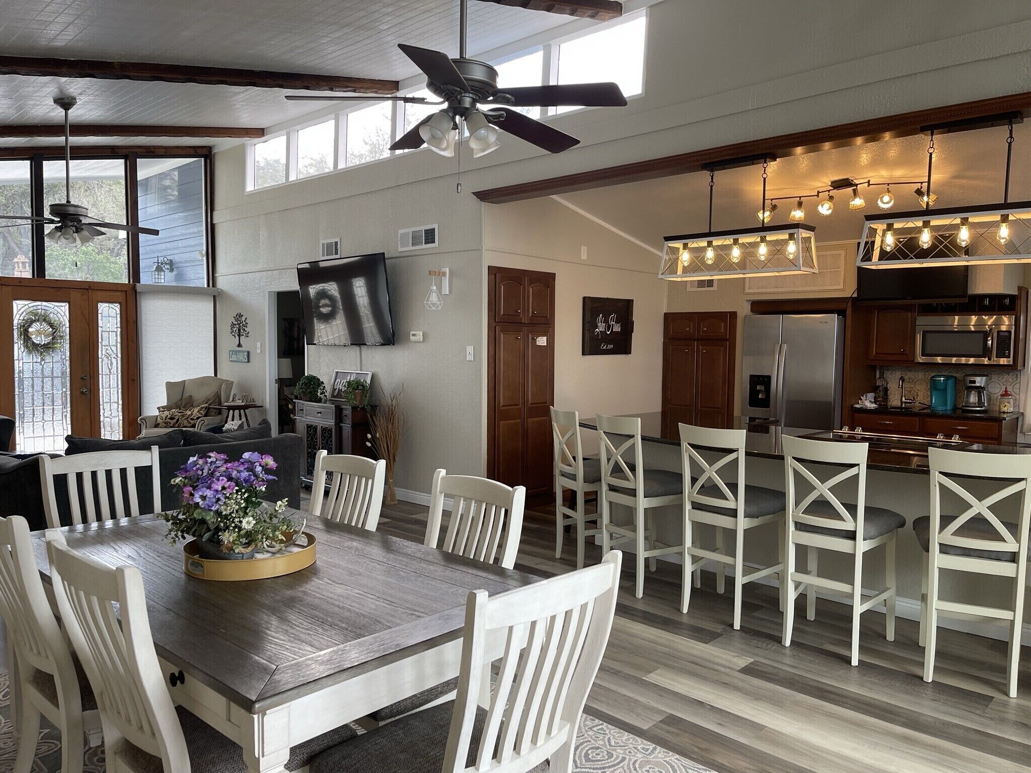 Un grand espace à manger avec une table en bois et des chaises blanches, une cuisine équipée d’appareils électroménagers modernes et un ventilateur de plafond.