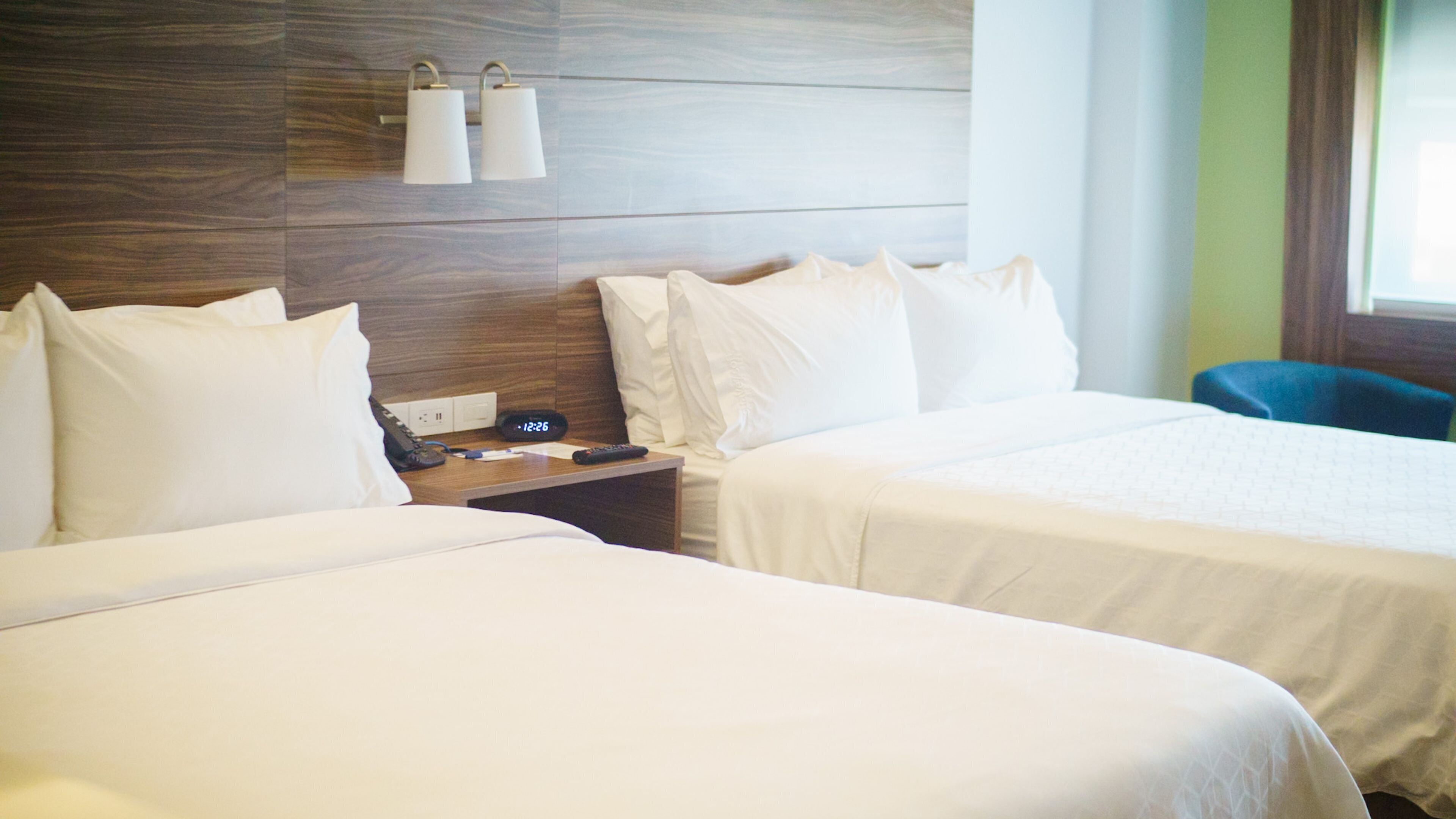A hotel room with two beds, a nightstand with a phone and alarm clock, and a window with curtains.
