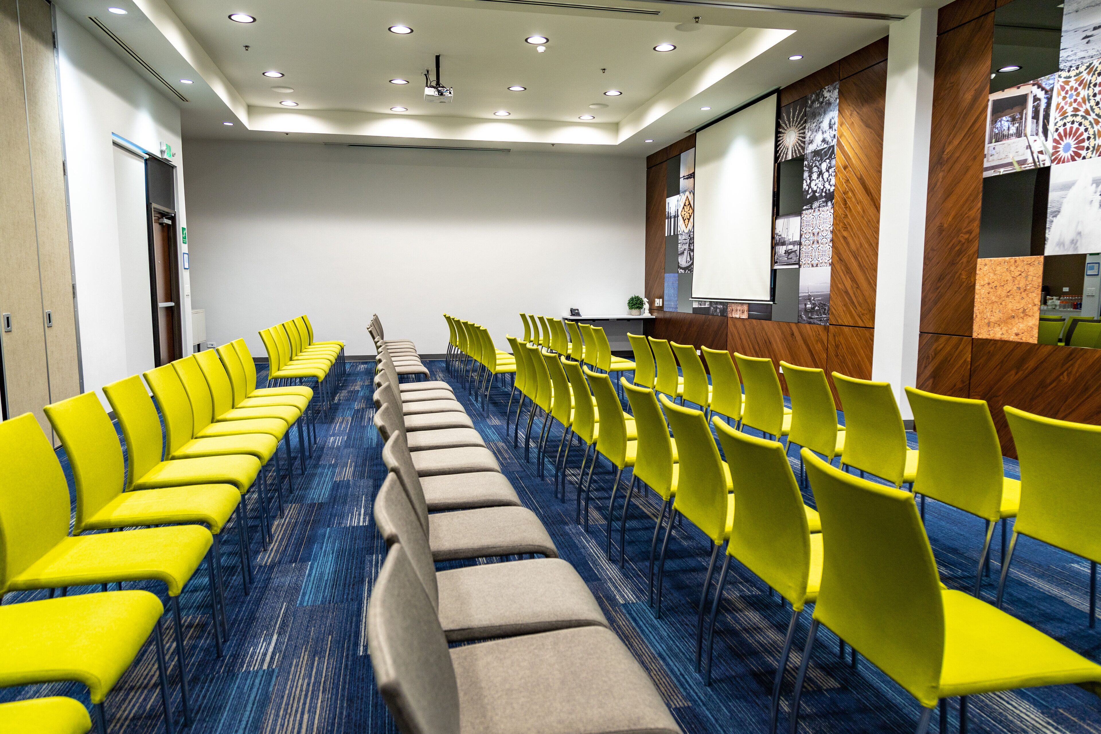 A conference room with rows of yellow and beige chairs, a wooden reception desk, and a blue carpeted floor.