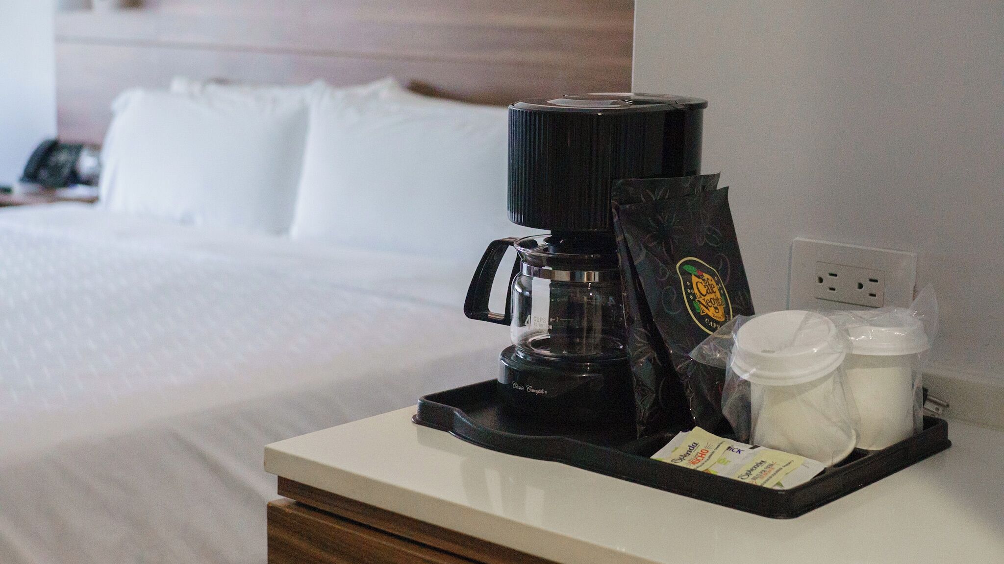 A coffee maker, a bag of coffee, and two containers on a hotel room's bedside table.