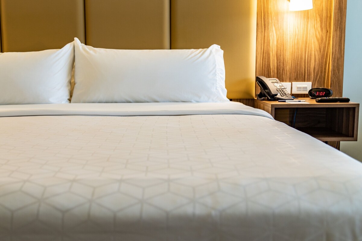 A neatly made bed with a geometric patterned bedspread, a bedside table with a lamp, a phone, and a digital clock displaying 3:02.