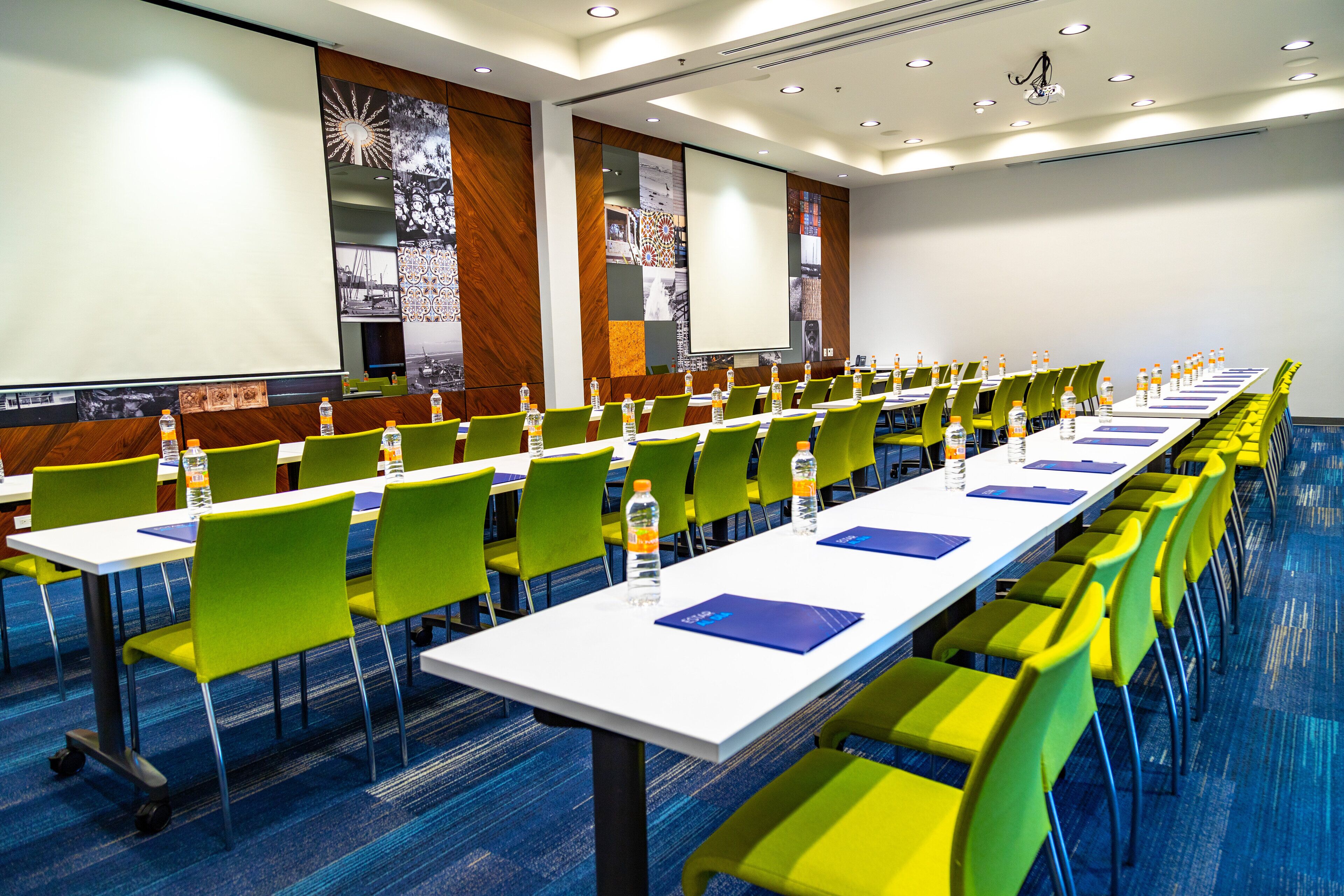 Une salle de conférence avec des rangées de chaises vertes et jaunes, des tables blanches et un grand écran à l’avant.