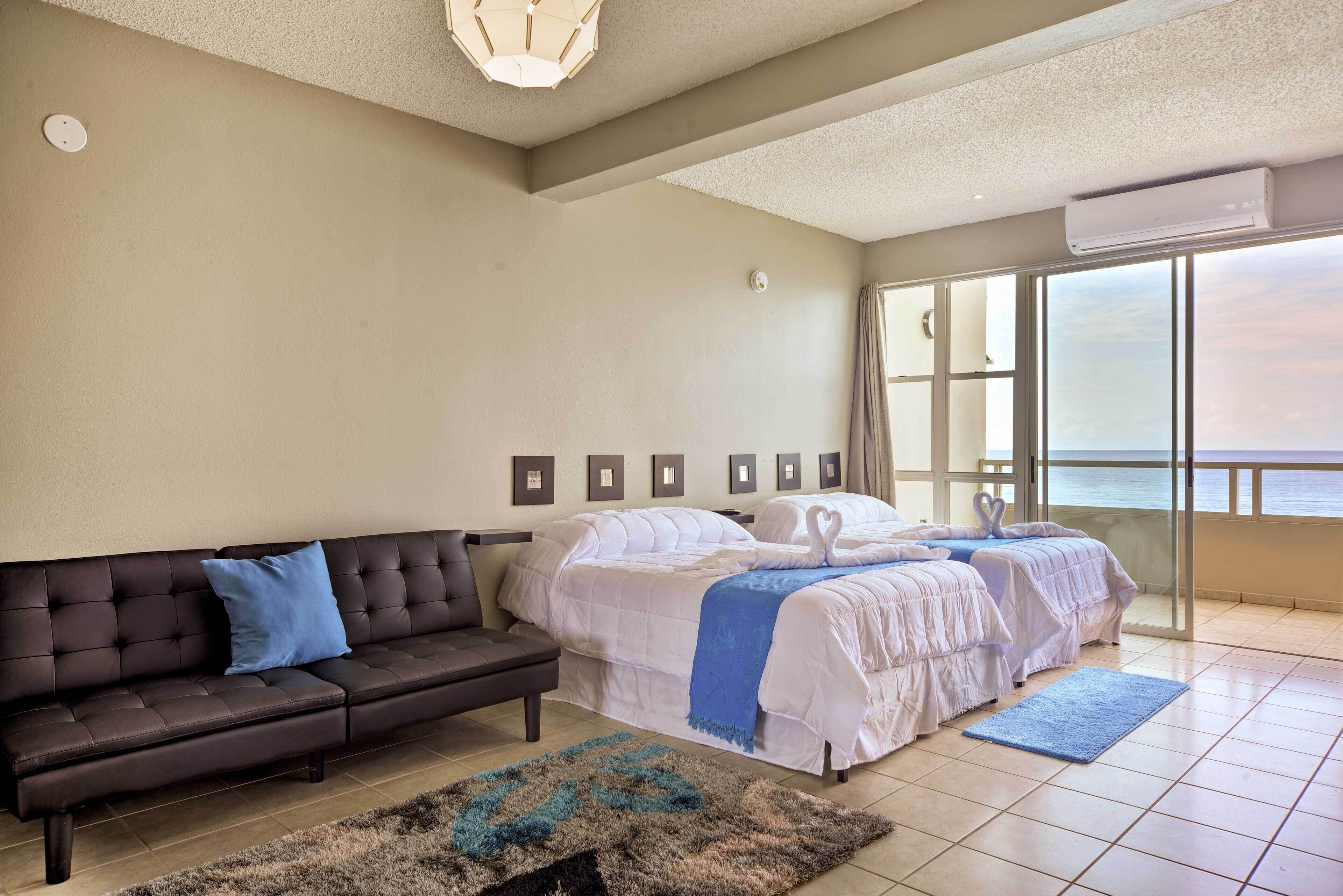 Quarto de hotel com uma cama grande, um sofá, um tapete e vista para o mar através de amplas janelas.