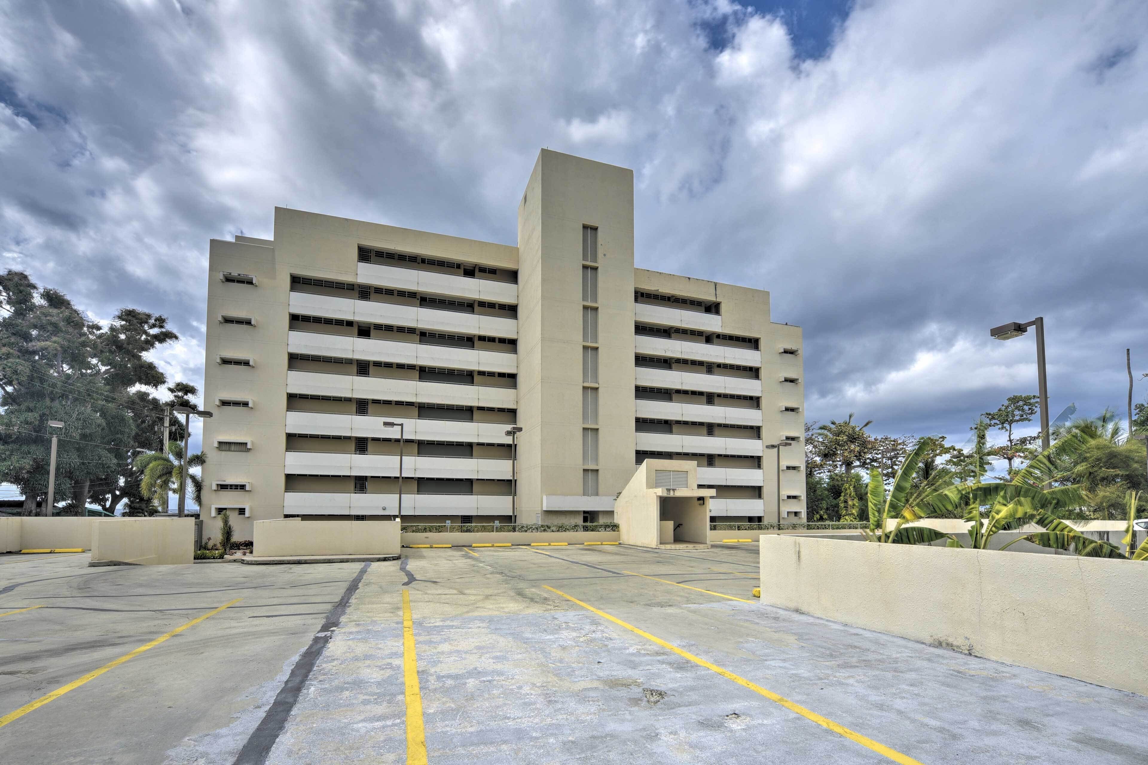 Un parking à étages doté d’une structure verticale centrale, entouré de verdure et surplombé par un ciel nuageux.