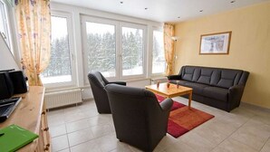 En stue med sofa, to stole, sofabord, radiator og store vinduer med udsigt til sneklædte træer.