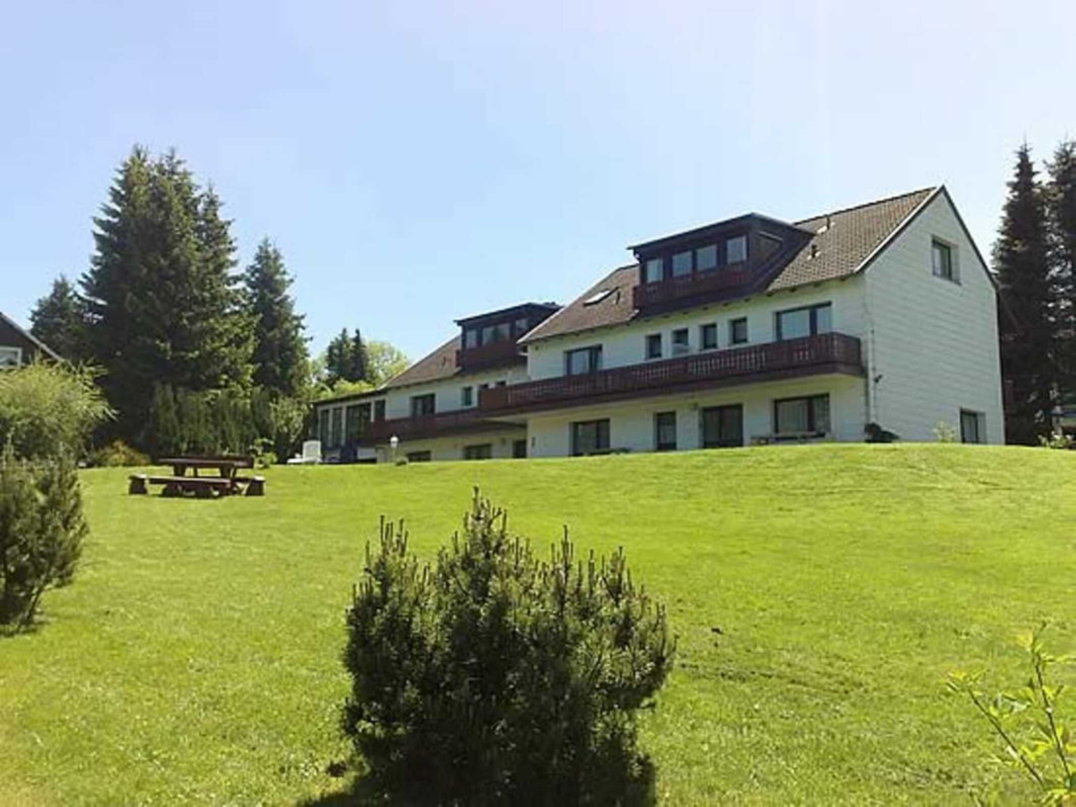 Ein zweigeschossiges Gebäude mit flachem Dach, einem Balkon im zweiten Stock und einer begrünten Rasenfläche davor.