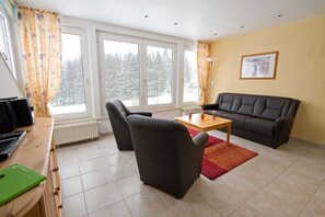 A living room with a sofa, two chairs, a coffee table, a radiator, and large windows with a view of snow-covered trees.