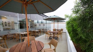 Un espace patio extérieur avec des tables rondes en bois et des chaises en osier, entouré de murs de verre et agrémenté d’un bac à fleurs blanc contenant un feuillage verdoyant.