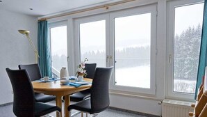Une pièce avec une table et quatre chaises, de grandes fenêtres donnant sur un paysage enneigé, un radiateur et une lampe.