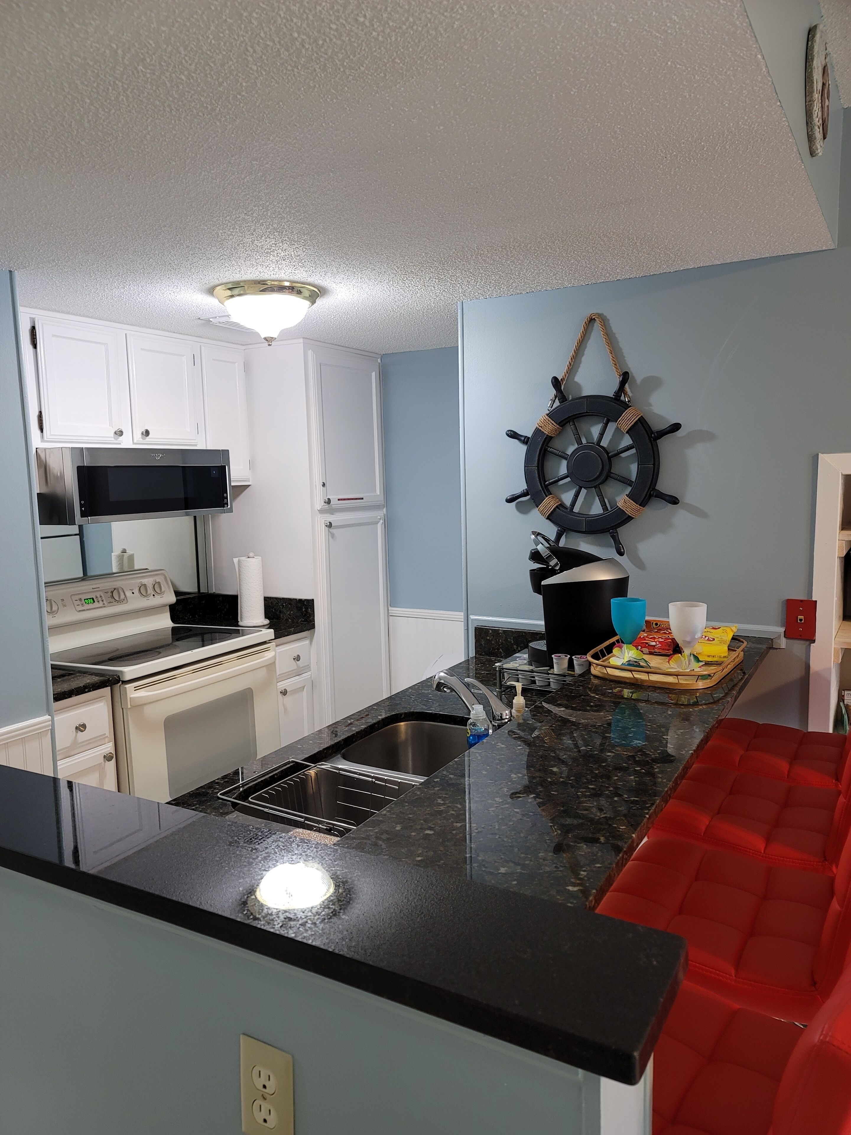 A kitchen with white cabinets, a stainless steel sink, a microwave, and a stove. There is a ship's wheel decoration on the wall and a red cushioned bench in the corner.