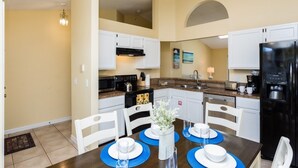 Cocina compacta con armarios blancos, un refrigerador negro y una mesa de comedor con individuales azules y platos blancos.