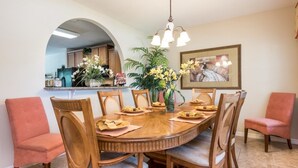 Un coin repas avec une table ronde en bois, six chaises et un lustre. On aperçoit une cuisine à travers une arche en arrière-plan.