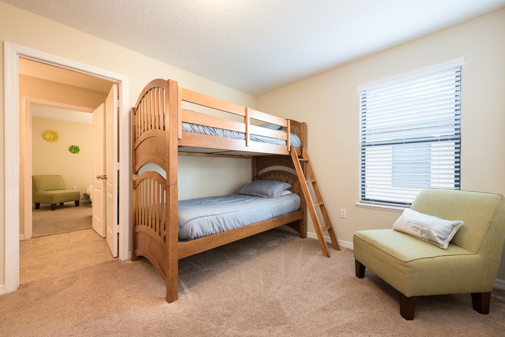 Una cama litera en una habitación con paredes color beige, una ventana con persianas y un sillón verde.