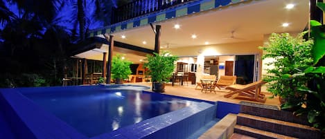 Relax by the pool surrounded by lush greenery and palm trees, with a covered patio offering a dining area and comfortable seating.