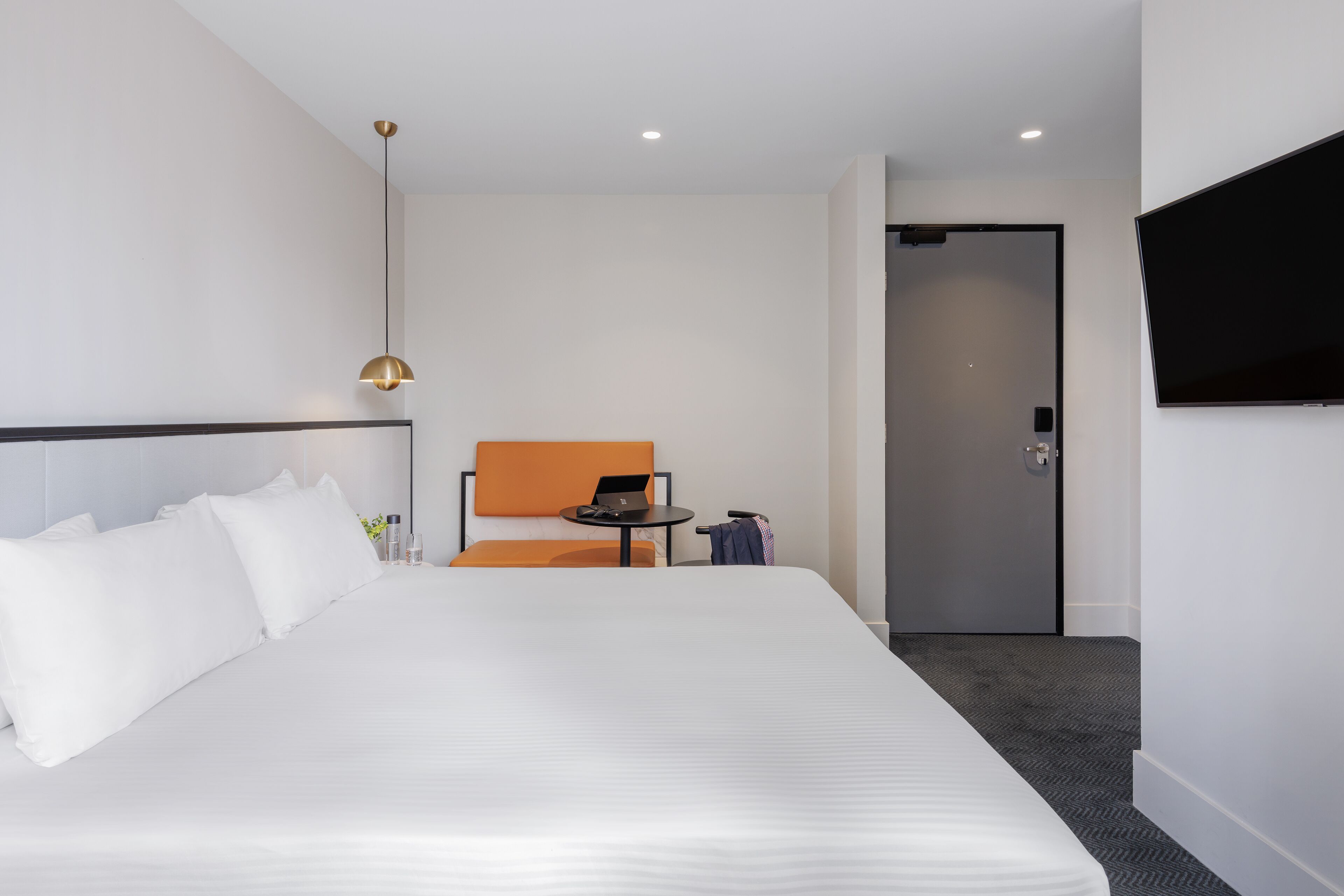 A hotel room with a large bed, a small table with a laptop, an orange chair, and a TV mounted on the wall.