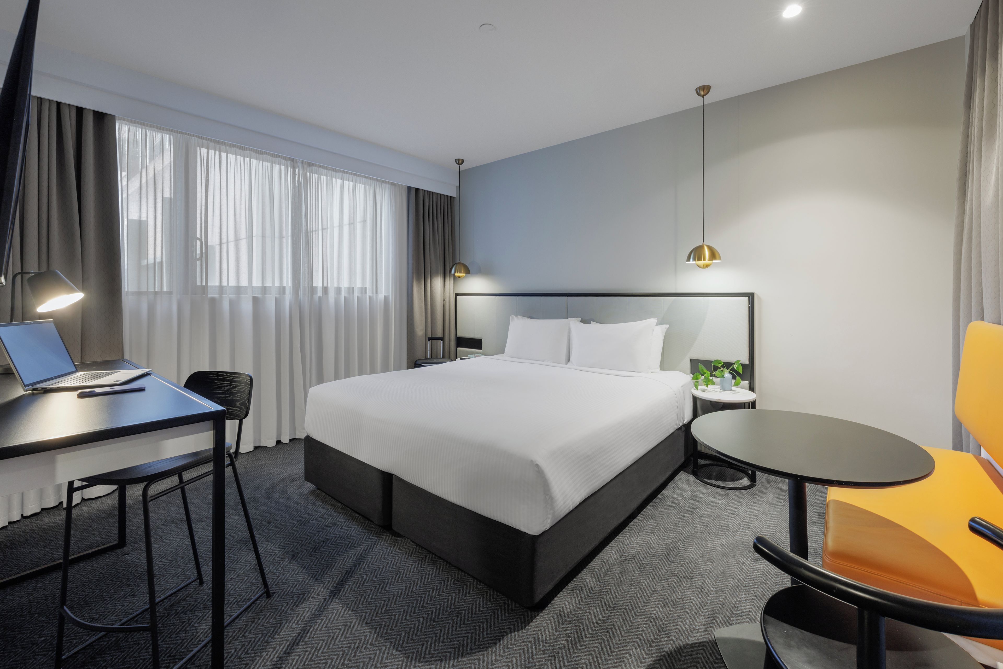 A hotel room with a bed, a desk with a laptop, a chair, a small table, and a lamp.