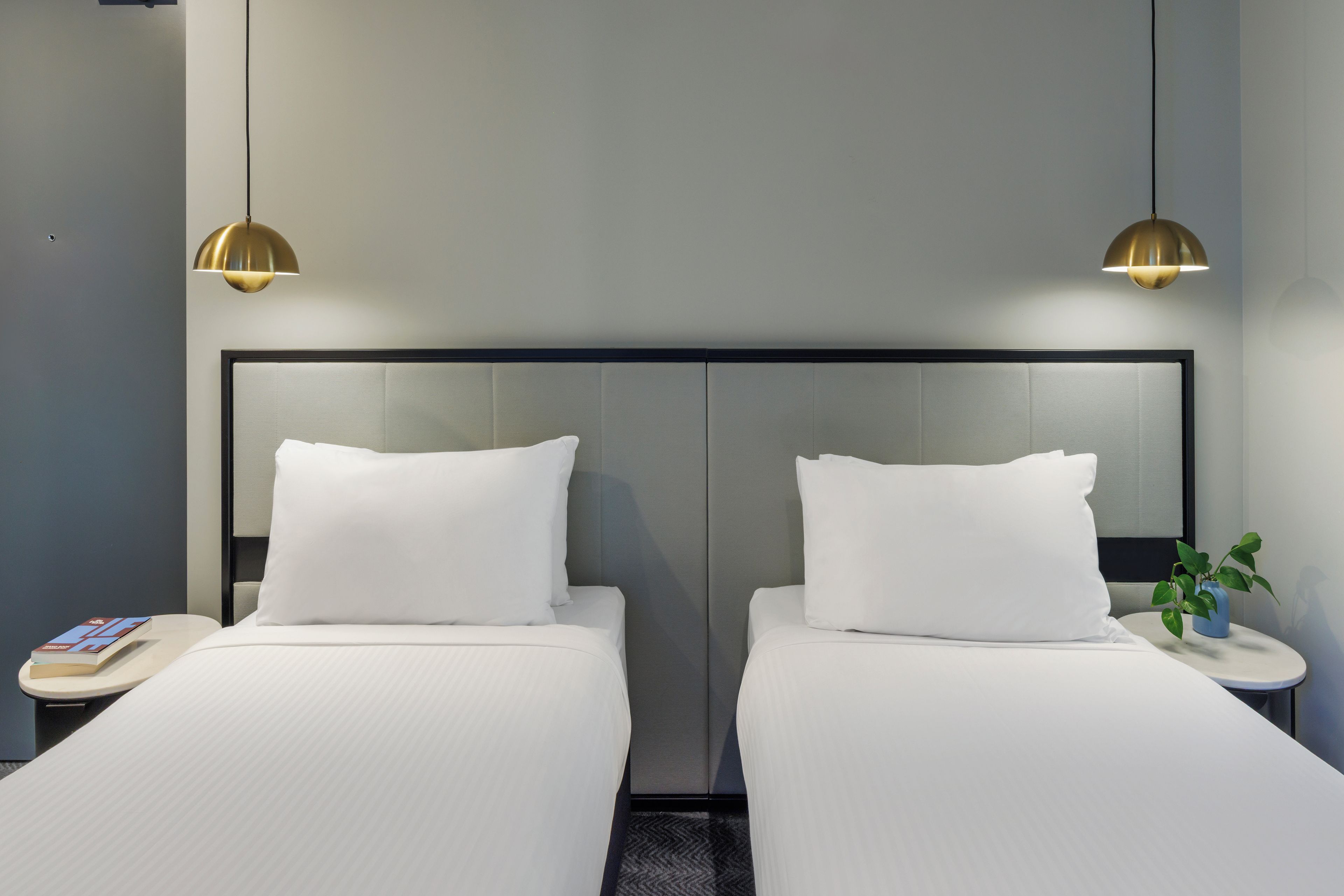 Une chambre d’hôtel avec deux lits, une table de chevet sur laquelle se trouve un livre et une petite plante, ainsi que deux lampes suspendues.