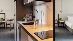 A modern kitchen with a built-in stove, a sink, and a countertop. There is a book on the counter and a glass of water. The room has a bed and a small table with a plant. - Brady Hotels Jones Lane (Melbourne)