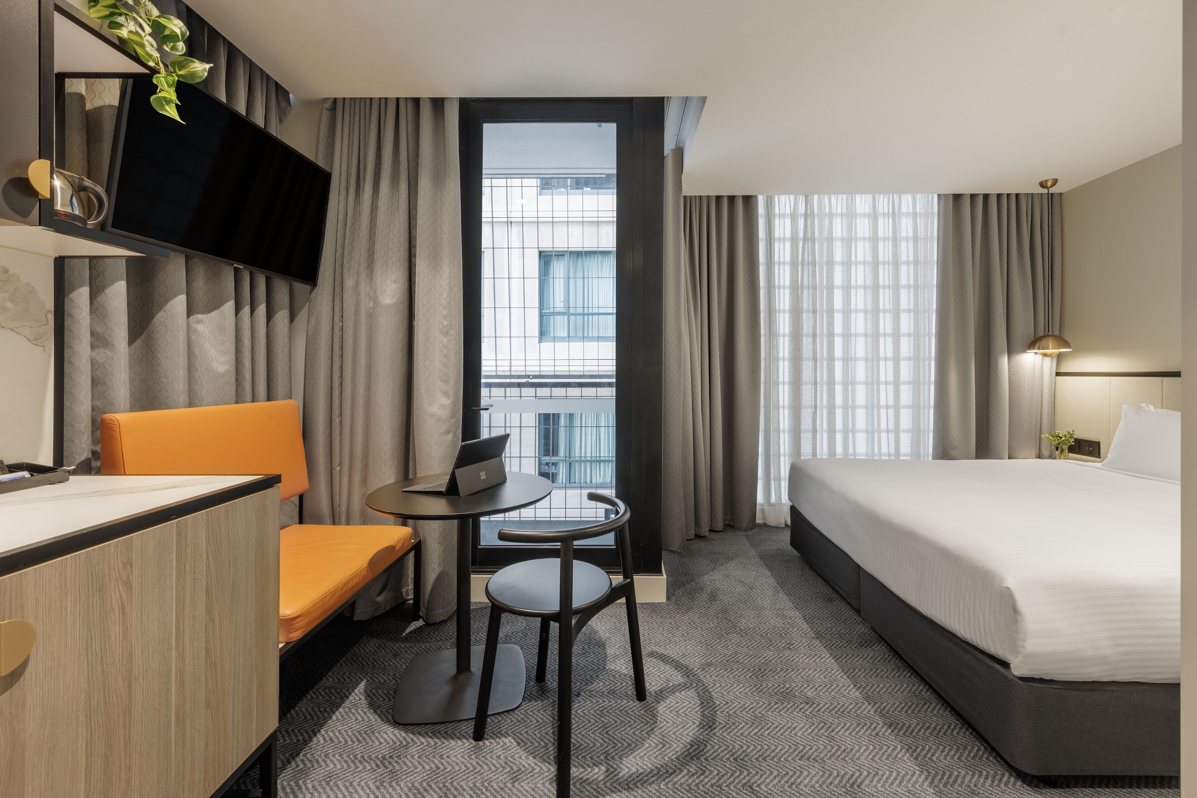 Habitación de hotel con cama, escritorio con computadora portátil, silla, televisor montado en la pared y vista al exterior a través de una ventana.
