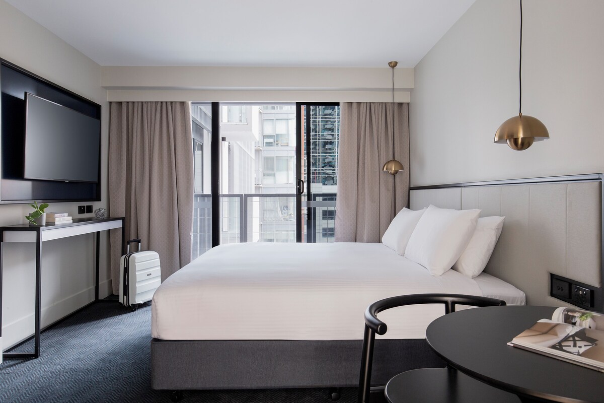 A modern hotel room with a large bed, a desk with a flat-screen TV, a chair, and a dining table.