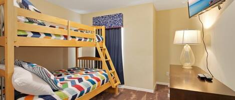 A bunk bed room with a TV on the wall, a desk with a lamp, and a window with curtains.