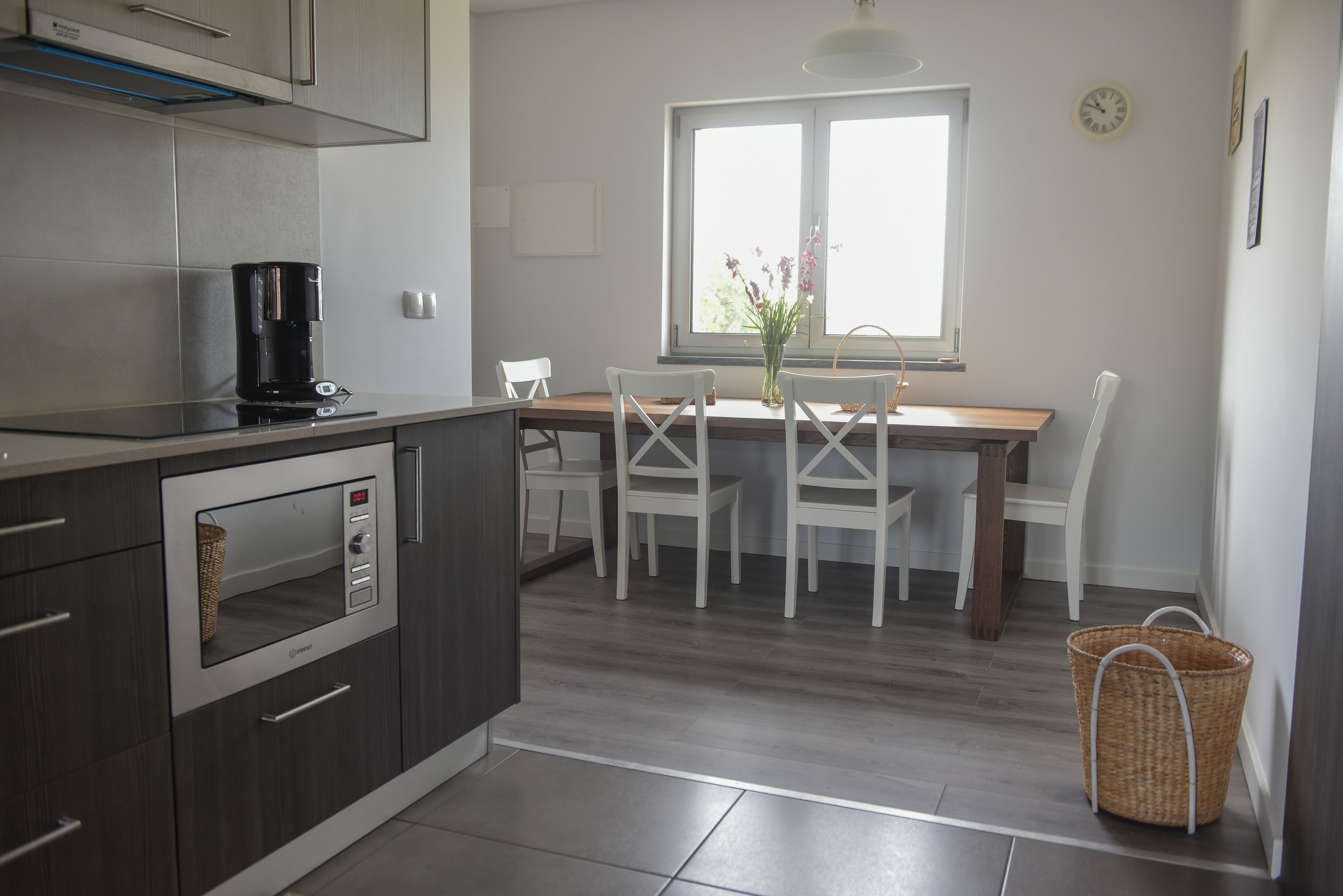 Una cocina moderna con horno empotrado, cafetera y zona de comedor con sillas blancas y mesa de madera.