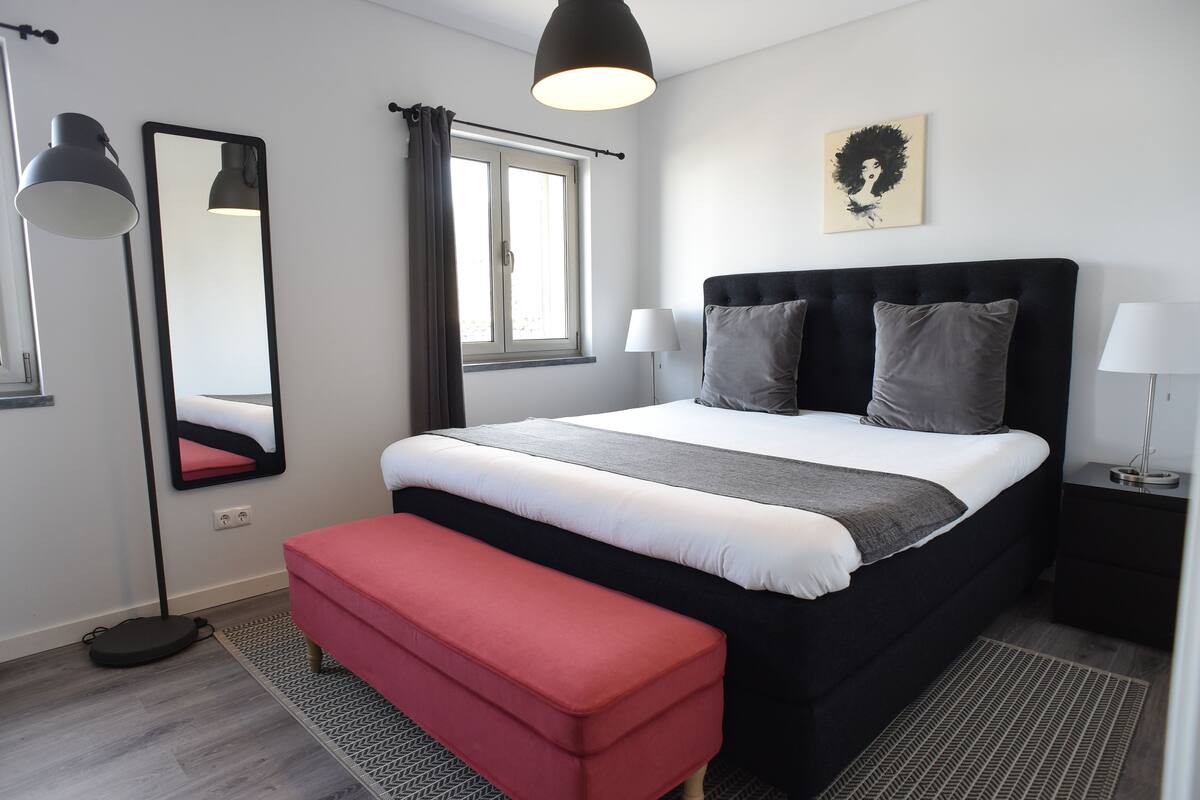 Un dormitorio moderno con una cama grande, un banco rojo, una lámpara de pie y una ventana con cortinas.