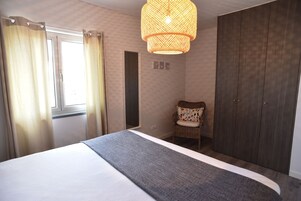 Un dormitorio con cama, una silla de mimbre, un armario y una lámpara colgante.