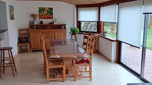 A dining area with a wooden table and chairs, a kitchen area with cabinets and a stove, and large windows offering a view of a garden. - Summer season, near Perros Guirec and pink granite coast, house 6 people (Louannec)
