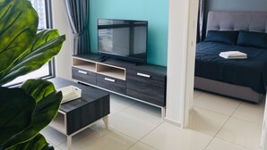 Une chambre d’hôtel moderne dotée d’un téléviseur à écran plat posé sur un meuble télé en bois foncé, d’un lit avec une literie sombre et d’une grande fenêtre avec des rideaux.