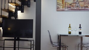Un salón moderno con un televisor de pantalla plana, una mesa de vidrio con botellas de vino y copas, y un cuadro de un paisaje urbano en la pared.