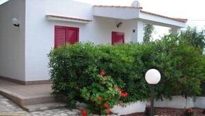 A white building with red shutters, a stone-paved entrance, and a well-maintained garden with red flowers.