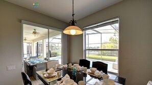A dining area with a glass table set for a meal, a ceiling fan, and large windows with a view of the outdoors. - Windsor Hills 2nd Premium 5/5 Lake View Home w/South Pool-Near Disney (Kissimmee)