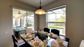Een eethoek met een glazen tafel, gedekt voor een maaltijd, een plafondventilator en grote ramen met uitzicht op de omgeving.