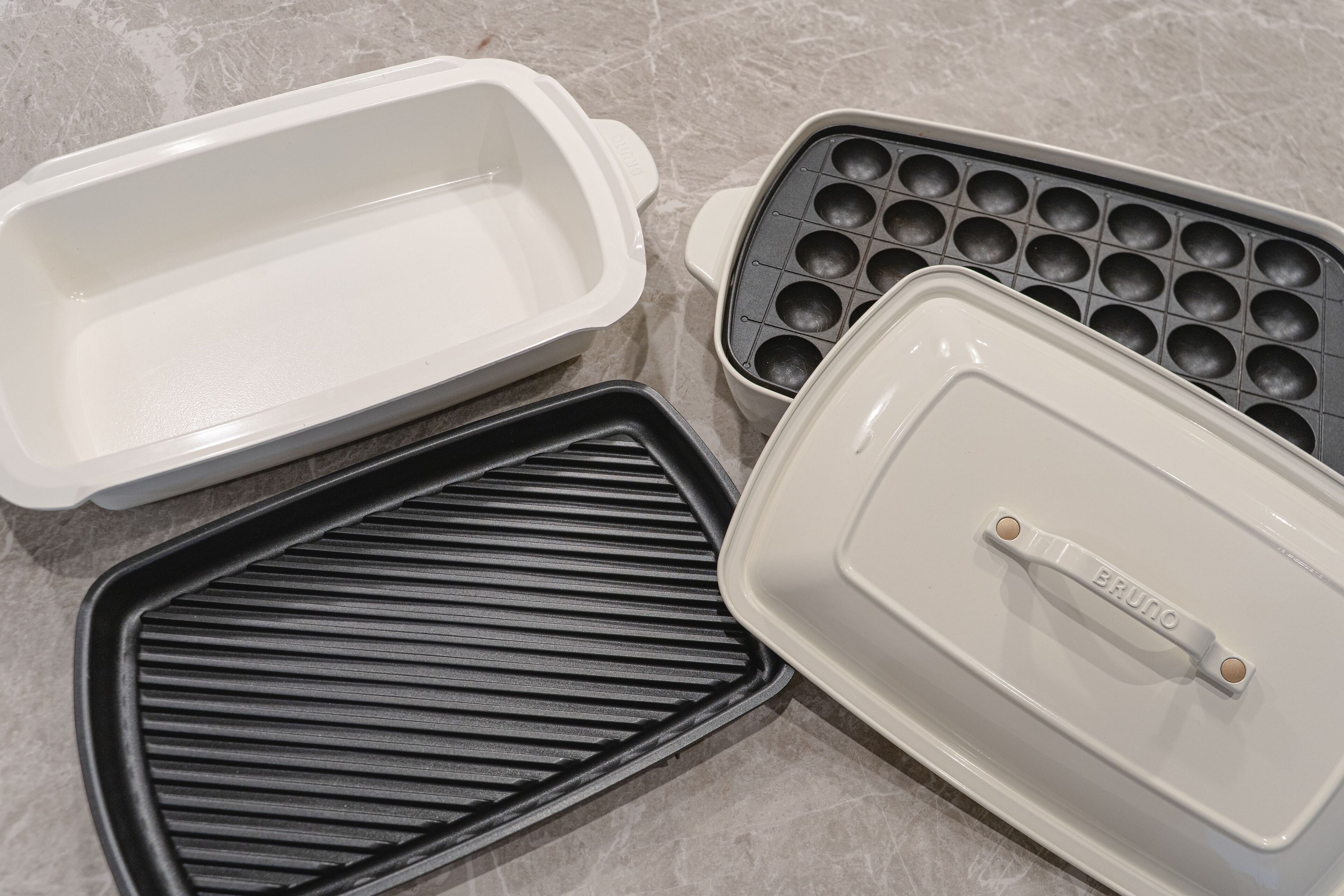 Four kitchen appliances: a white rectangular container, a black grill pan, a white muffin tray, and a white container with a handle labeled "BRUNO".