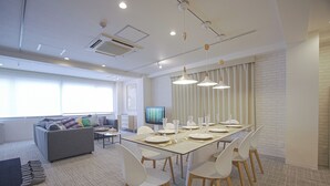 A modern office space with a large dining table set for six, white chairs, a gray sofa with colorful cushions, and a large window with blinds. - INOVE VILLA OSAKA (Osaka)