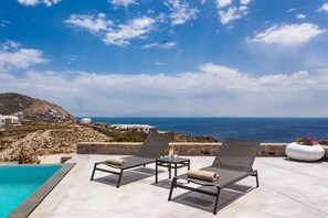 Un espace aménagé au bord de la piscine, avec deux chaises longues, une petite table sur laquelle se trouve un verre, et une vue sur la mer et les bâtiments situés au loin.