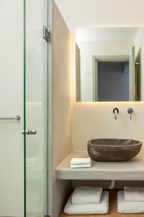 Une salle de bain avec une porte de douche en verre, un grand miroir, un lavabo en pierre et des serviettes soigneusement pliées, rangées sur une étagère.