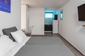 Une chambre d’hôtel moderne avec un lit, une chaise, une petite table, un téléviseur et, au fond, une kitchenette.
