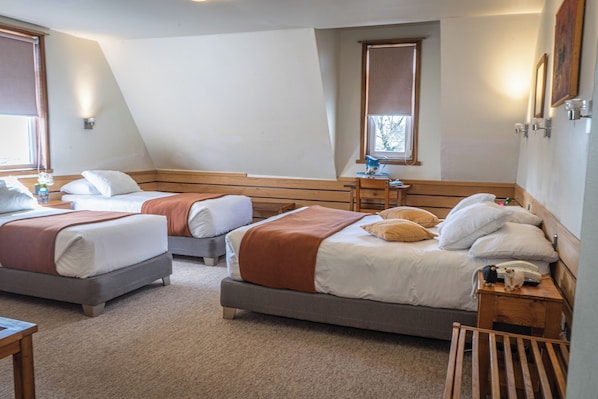 A hotel room with two beds, a wooden headboard, a bedside table with a lamp, and a window with blinds. - Hotel Museo El Greco (Puerto Varas)