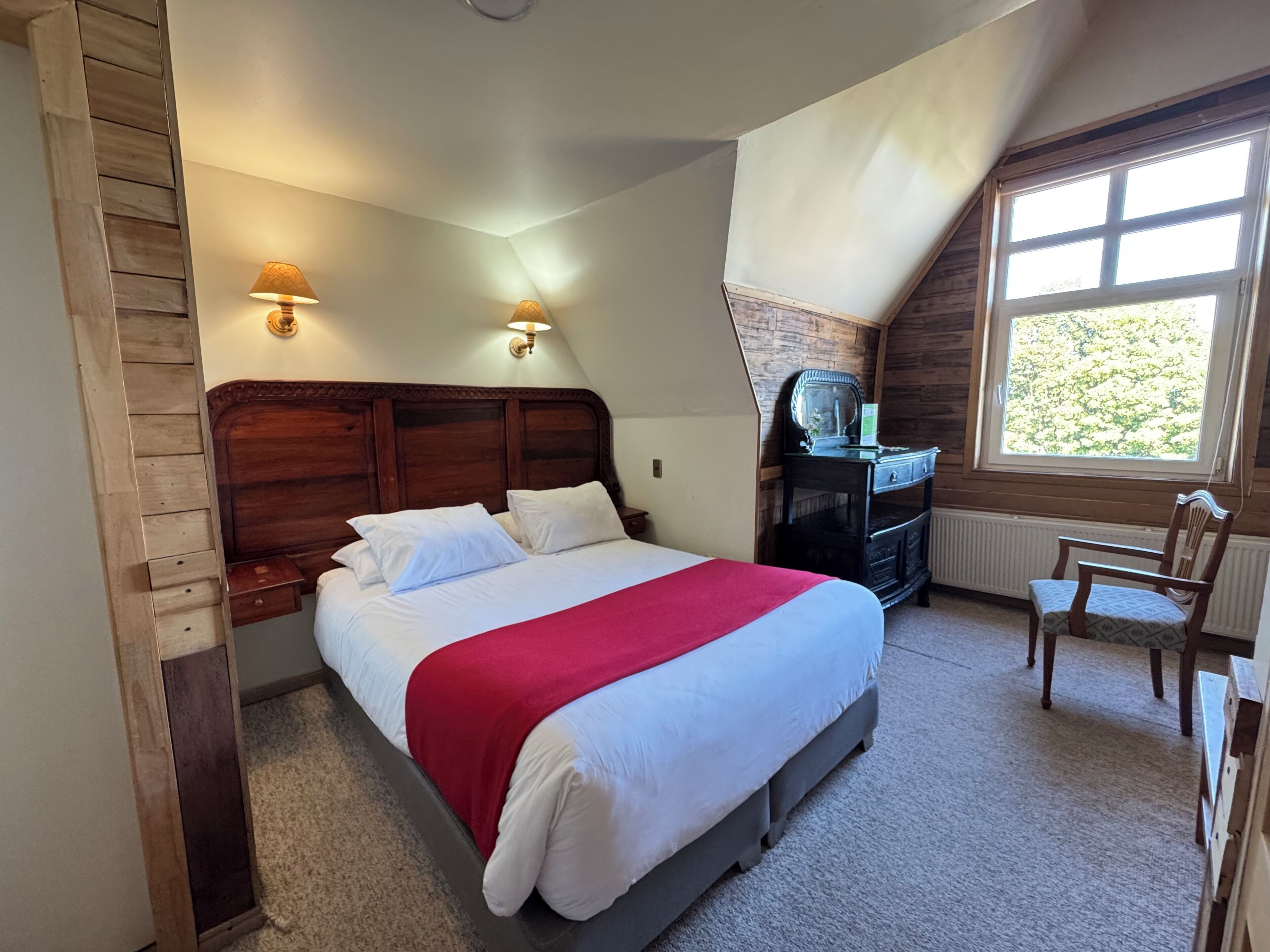 Un dormitorio con cabecera de madera, cama con ropa de cama blanca y manta roja, cómoda con espejo y ventana con vistas a árboles.