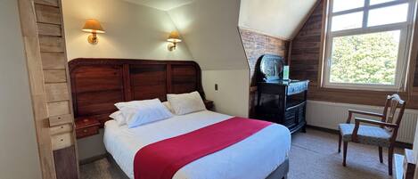 A bedroom with a wooden headboard, a bed with white bedding and a red blanket, a dresser with a mirror, and a window with a view of trees.
