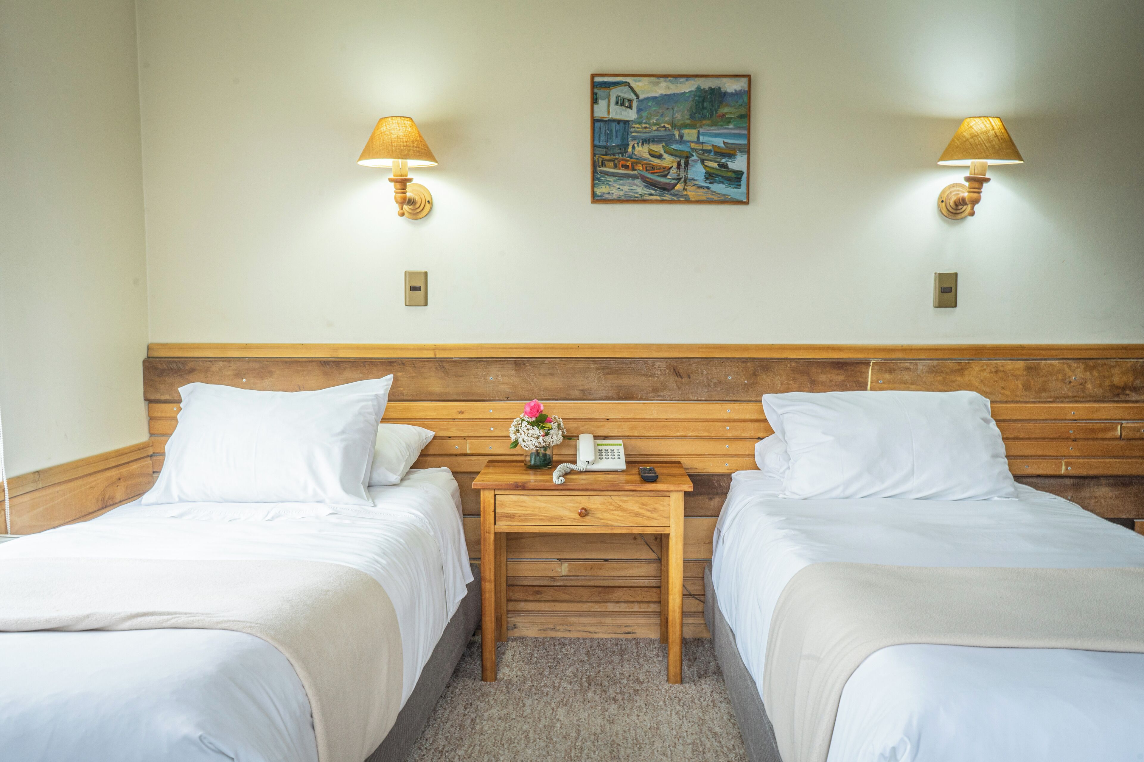 A hotel room with two beds, a wooden nightstand with a telephone and a vase of flowers, and two wall-mounted lamps.
