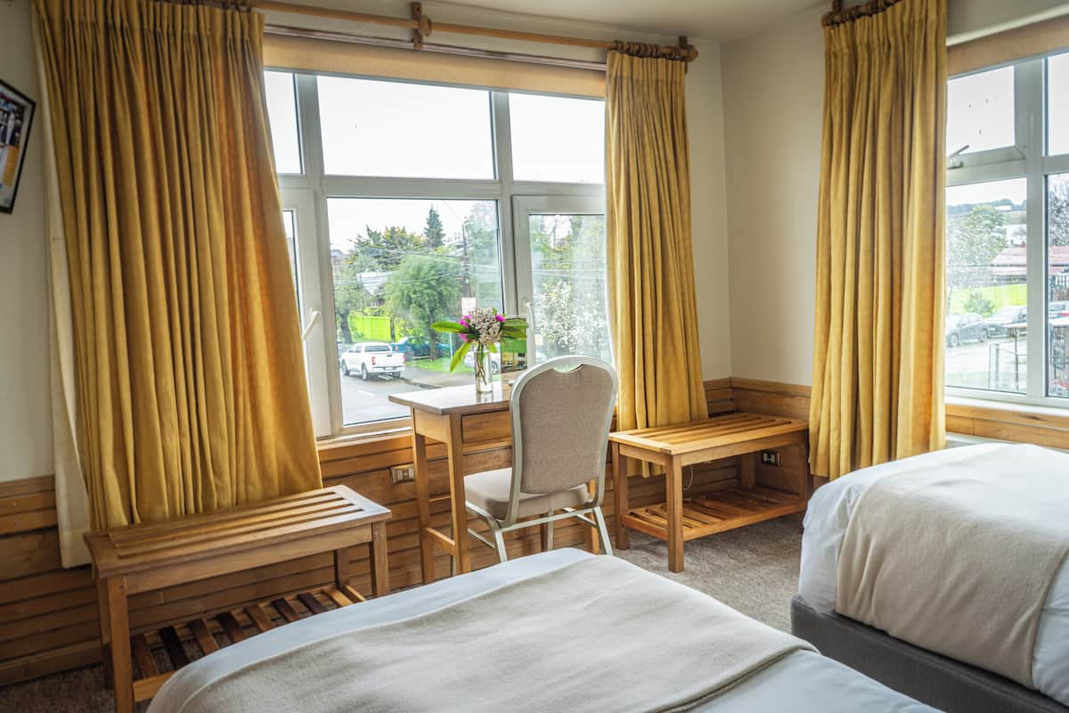 A hotel room with two beds, a desk, and a chair. There is a window with curtains, and a view of the outside.
