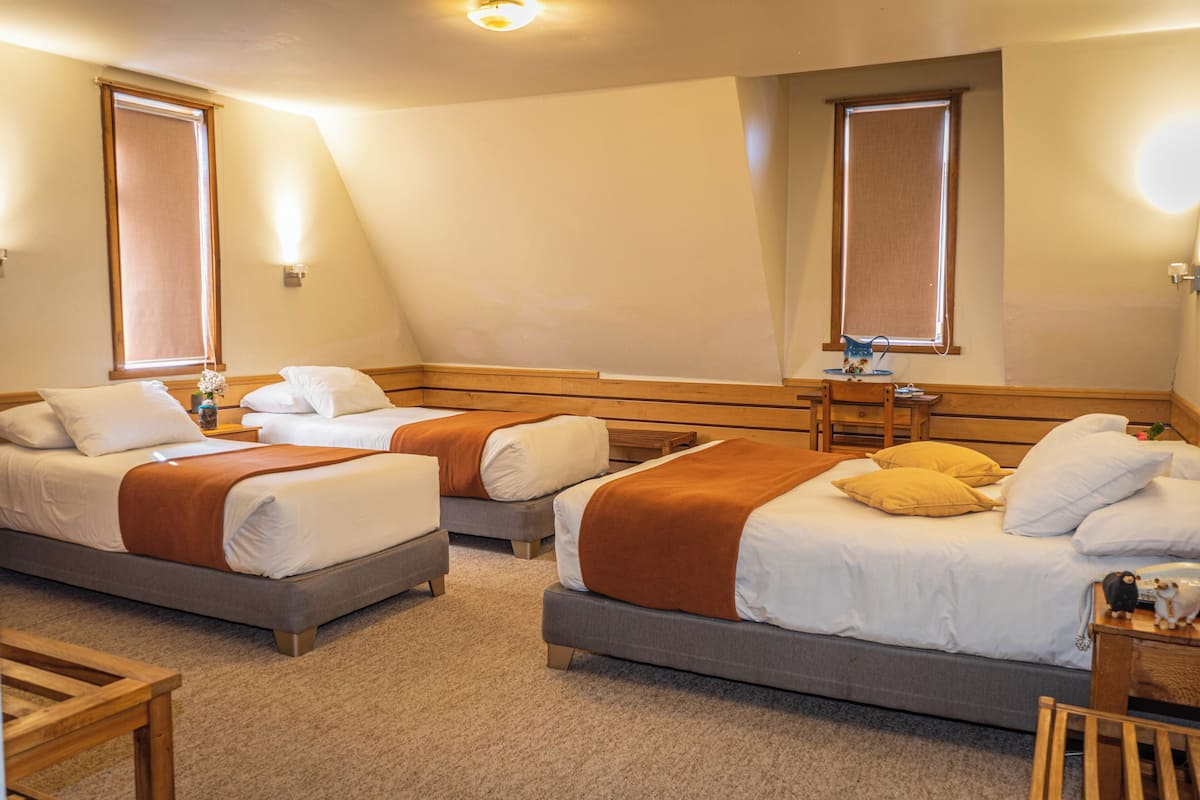 A hotel room with three beds, each with white bedding and brown throw blankets. There are wooden headboards and nightstands with lamps.